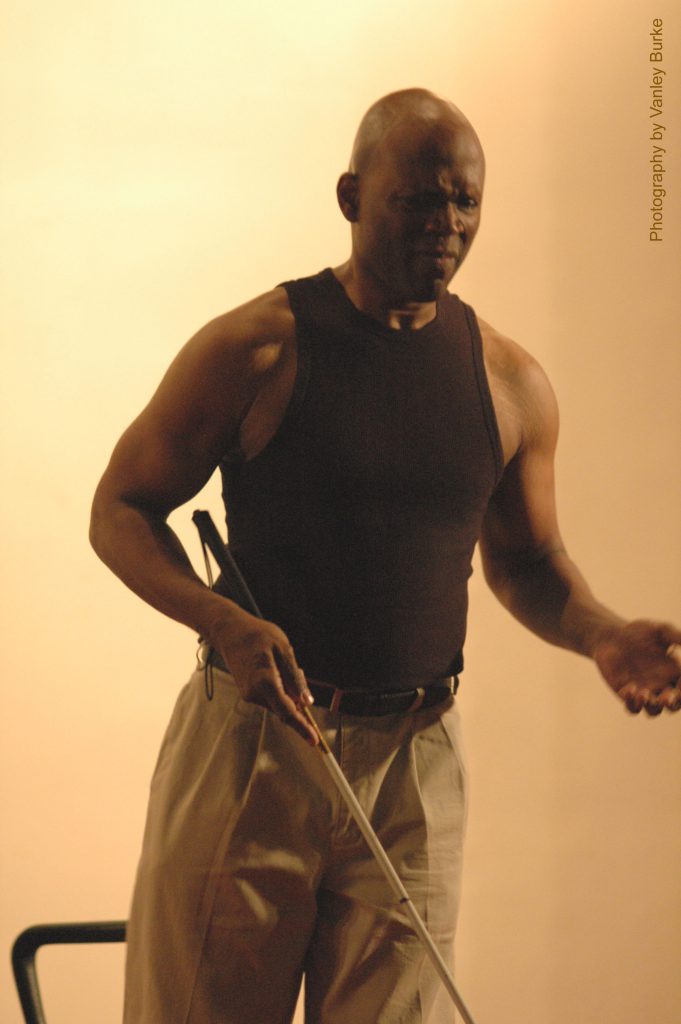 Lynn performs to camera in Weights holding out his white cane with one hand and gesticulating with the other.
