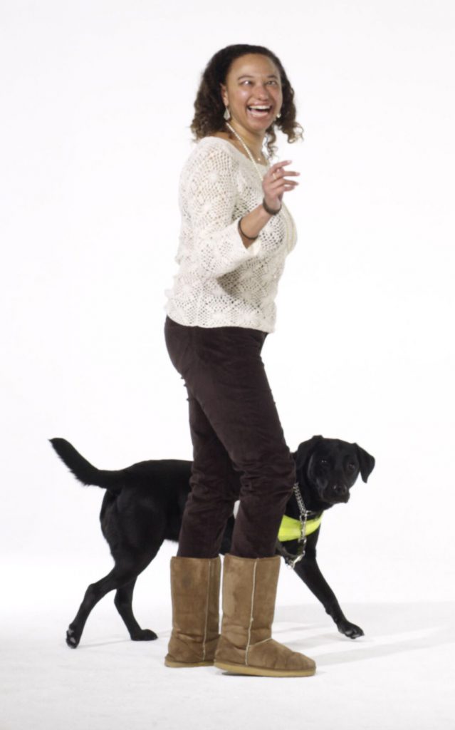 Maria stands and looks at the camera with an animated expression, joined by her black guide dog Penny