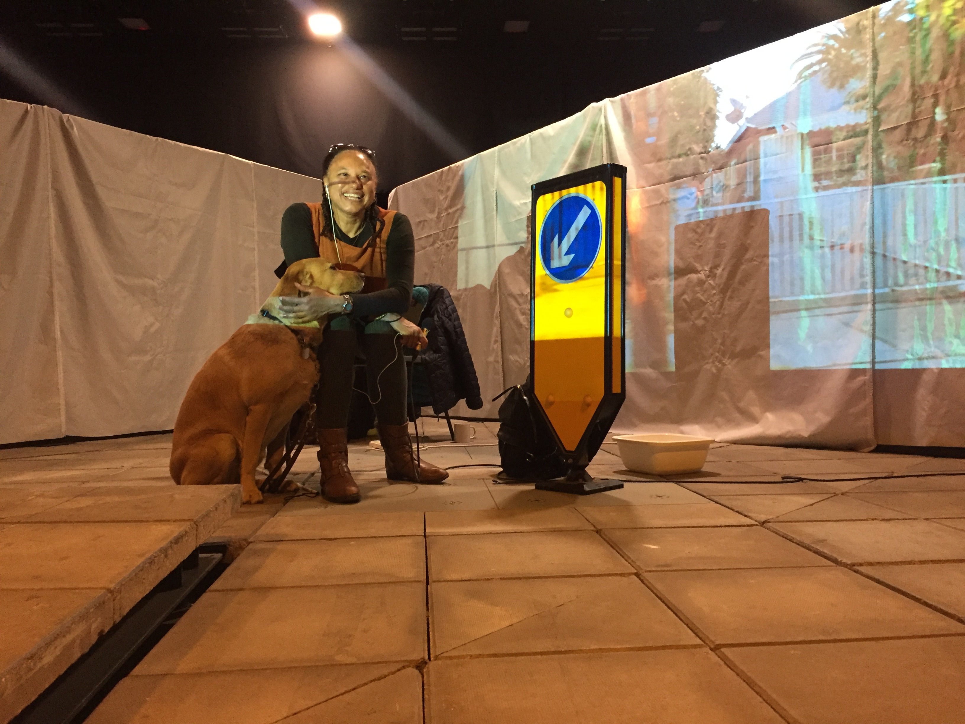 Maria sits between two screens smiling at camera and holding a mic. Her guide dog Bella and a road sign stand nearby