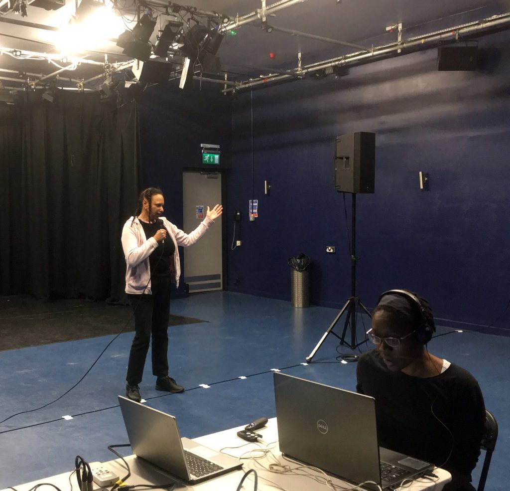 Maria standing in theatre studio  gestures while another woman of colour sits at a laptop