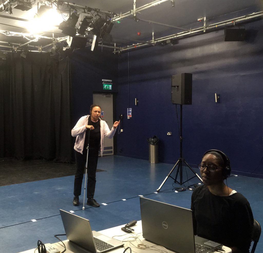 Maria standing in a studio space gestures with a white cane to another  black woman sitting at a laptop on a  table