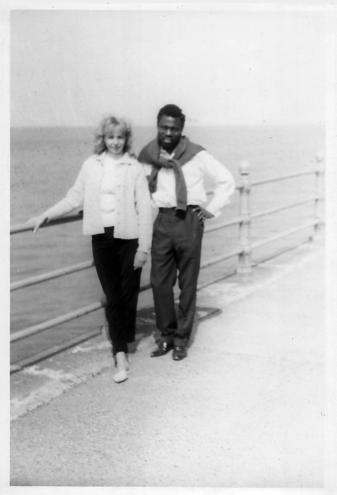 Young black man and white woman standing together by a rail on a seaside promonard smiling out at camera in fashions of the 1960’s