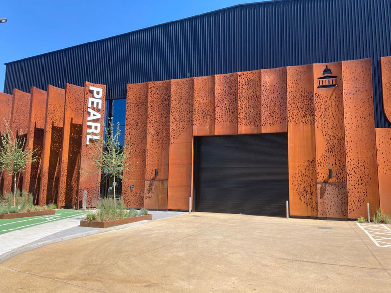 Front of a huge building with vertical black corrugated sheeting  running full height, and rusty bronze Corten steel panels covering the lower two thirds of the wall. Big sections are block cut out around black doors and massive letters P.E.A.R.L run vertically upwards  in white.