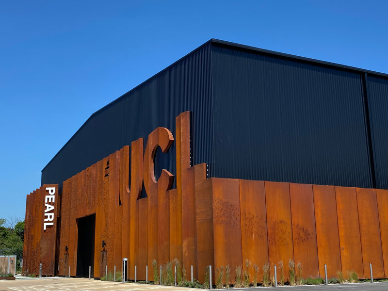 The approach to Pearl  from a distance and facing the right hand corner of the short end of this rectangle building, against a deep blue sky. The ‘L’ of UCL is closest in the steel rust-coloured wall.  On the left the name PEARL in white capital letters running vertically down from the top of the steel wall  and positioned to the left of the entrance door.