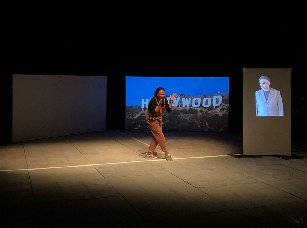 Maria gestures to Lynn on a down stage screen while the Hollywood sign appears large on the right hand screen behind
