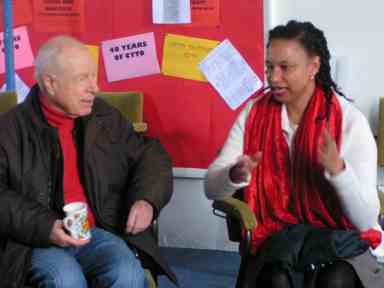 Maria, a biracial woman in a white jumper, sits talking next to Peter Brook, an elderly white man who is holding a cup of tea