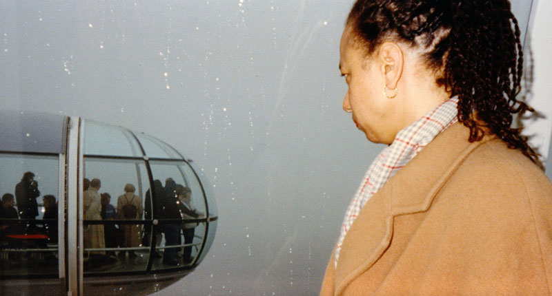 Maria in a coat stands in profile against a window speckled with rain, through which a pod of the London Eye can be seen in the distance
