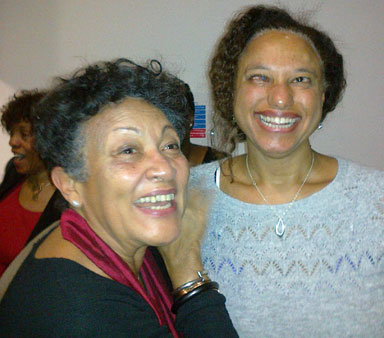 Maria in a pale blue jumper stands smiling with Yvonne Brewster, an older woman of African heritage wearing a black jumper