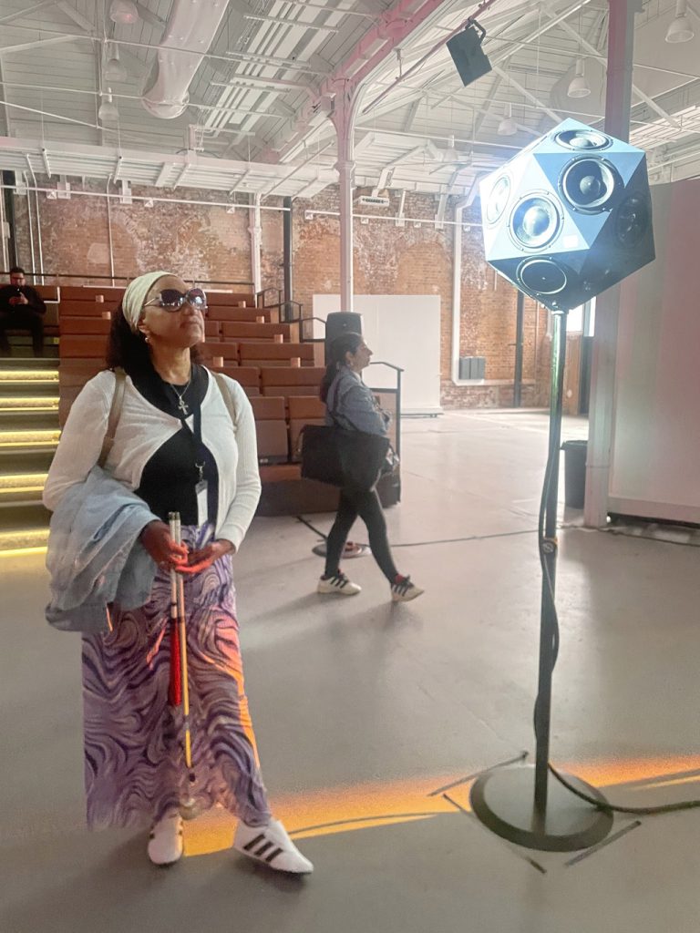 Maria  in a shaft of light looks up at  a 20 sided Iko speaker  above her on a stand.