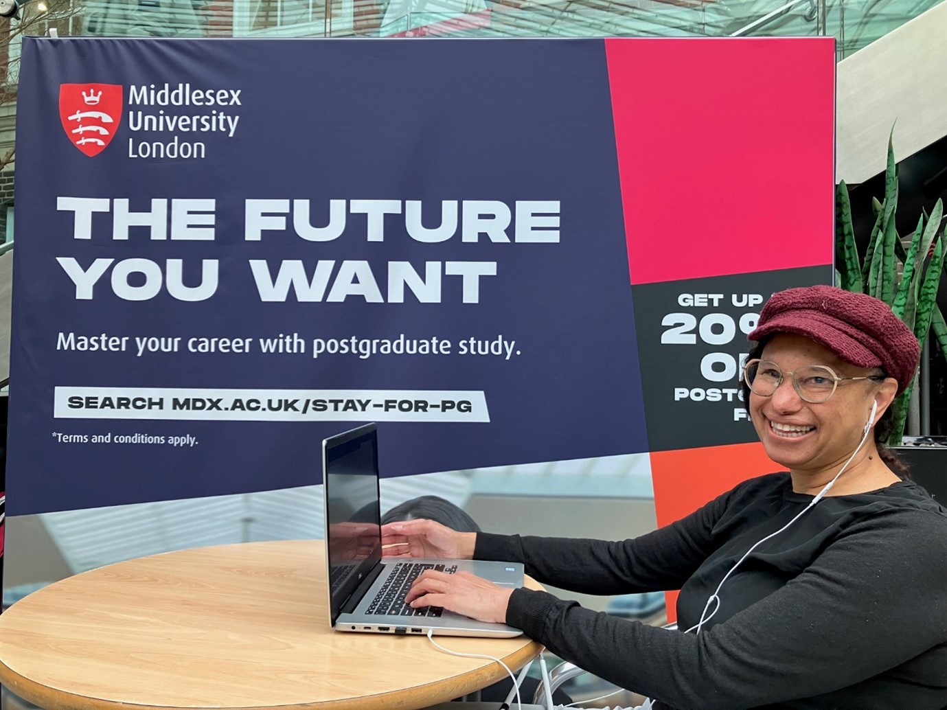 Maria sits with a laptop smiling and wearing a black top and Burgundy peaked cap next to a large Middlesex University banner