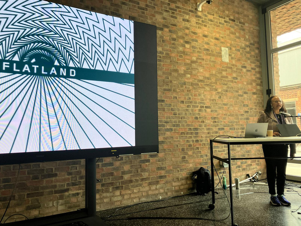 Maria wearing a pink and grey check blazer stands at a lectern delivering a paper  in front of a large black and white Flatland logo on screen
