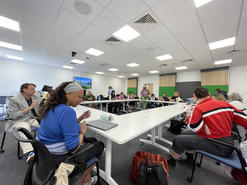 Maria sits at a large seminar table in front of a laptop addressing a group of  delegates.
